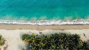 Eine Vogelperspektive zeigt einen Strand mit klarem türkisblauem Wasser, weißem Sand und dichter Palmenbepflanzung.