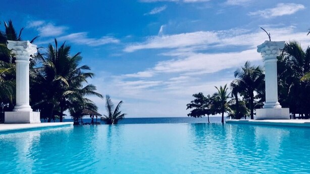 Ein klares, blaues Schwimmbecken mit weißen Säulen und Palmen, umgeben von wolkenlosem Himmel.