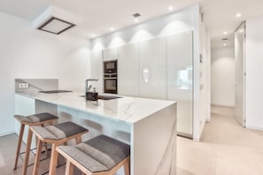 A modern kitchen with a marble countertop, a built-in oven, and a sink. There are three bar stools with cushioned seats.