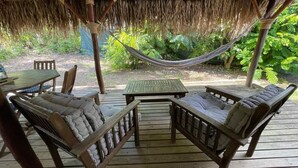 Una terraza de madera con techo de paja, dos sillas con cojines, una mesita y una hamaca.