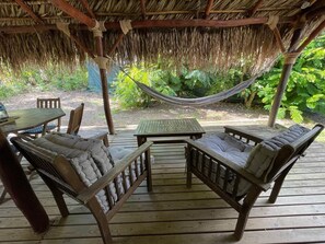 Une terrasse en bois surmontée d’un toit de chaume, deux fauteuils confortables, une petite table et un hamac.