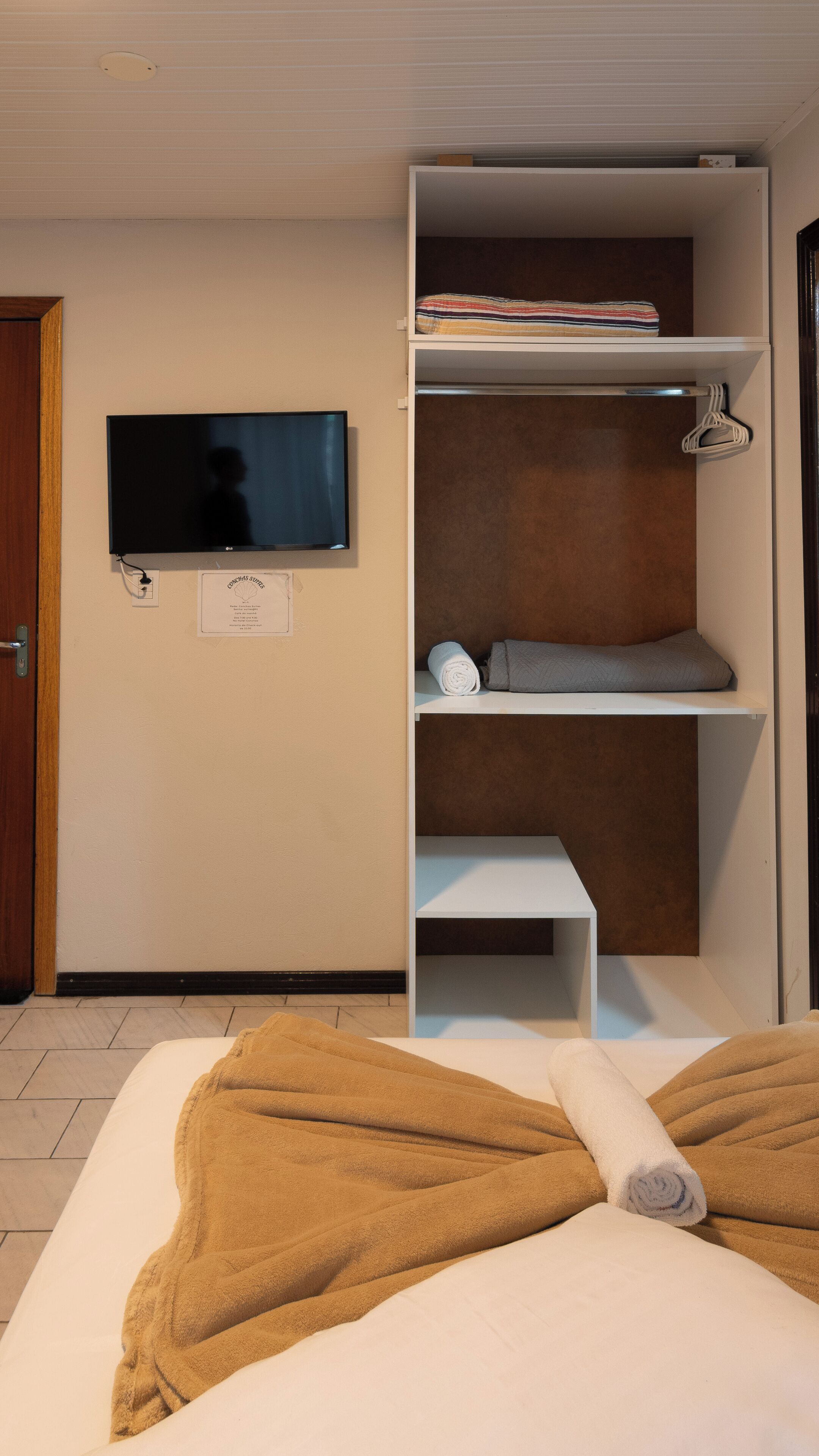 Une chambre d’hôtel comprenant un lit, un téléviseur fixé au mur, une armoire avec des étagères et une porte.