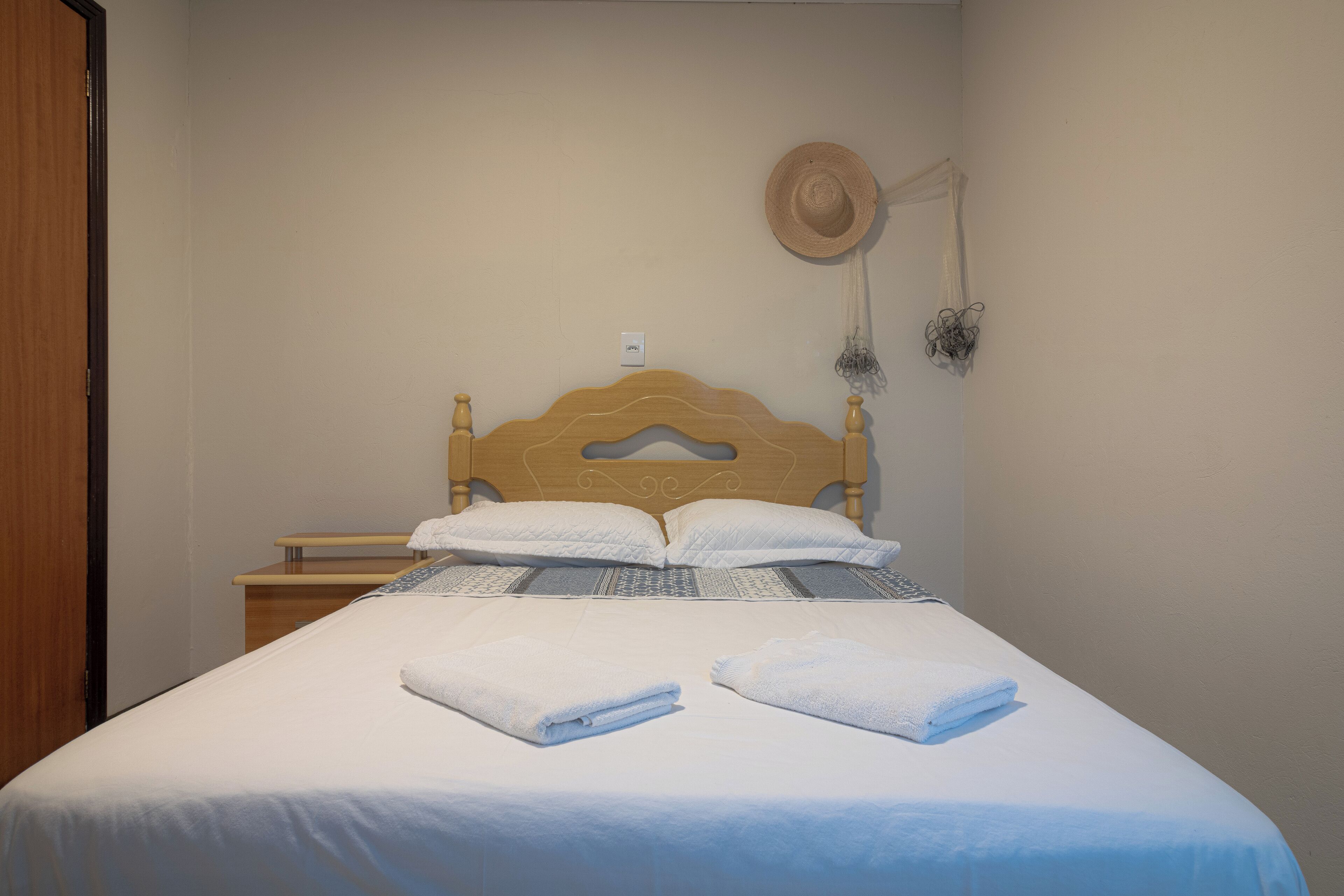 A neatly made bed with white linens and two folded towels, a wooden headboard, a small bedside table, and a wall-mounted hat and decorative item.