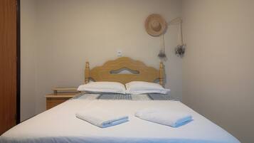 A neatly made bed with white linens and two folded towels, a wooden headboard, a small bedside table, and a wall-mounted hat and decorative item.