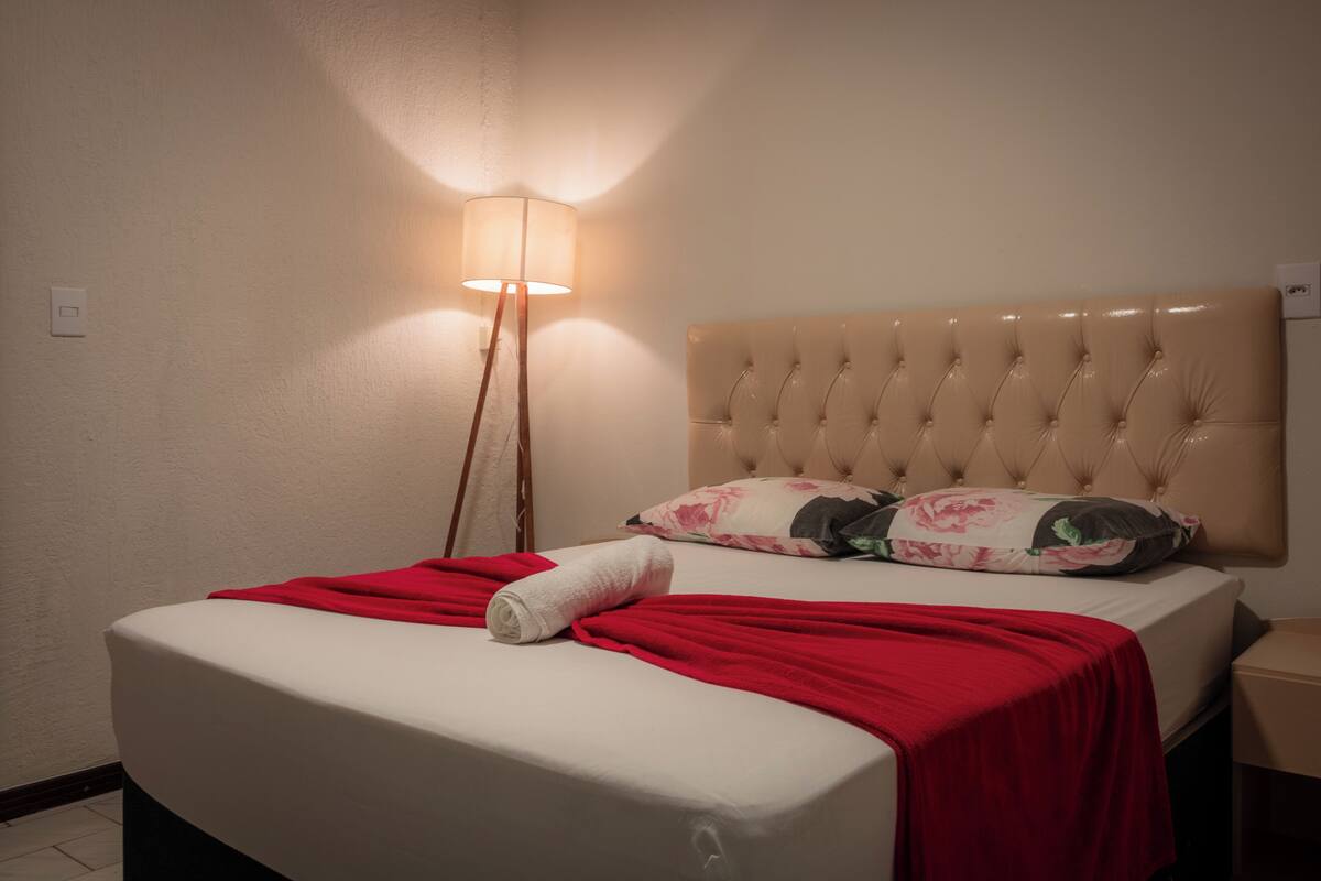 Una cama bien tendida con una manta roja y una toalla enrollada, un cabecero tapizado en color beige y una lámpara de pie que ilumina la pared.