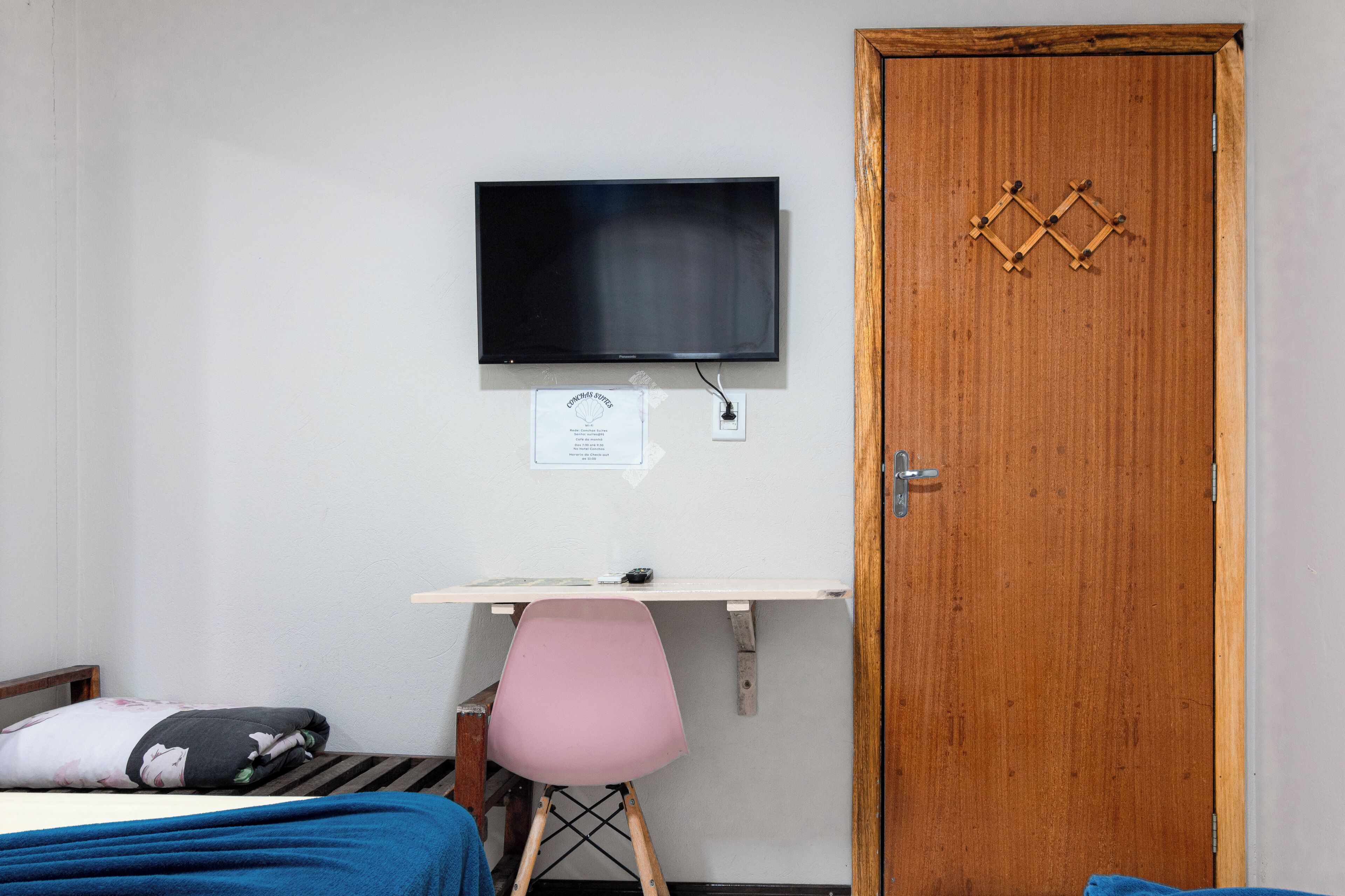 A bedroom with a bed, a desk with a chair, a television mounted on the wall, and a wooden door.