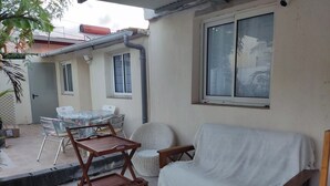 Eine Terrasse mit einer weißen Couch, Korbstühlen und einem Holzwagen. Ein Tisch mit Stühlen und ein Fenster mit Blick.
