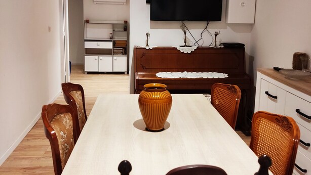 Ein Essbereich mit einem langen Tisch, Stühlen, einer Vase, einem Fernseher, einem Klavier und einer Küchenzeile.