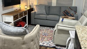 Ein Wohnzimmer mit Flachbildfernseher, grauem Sofa, Sessel und einem kleinen Tisch mit Lampe.