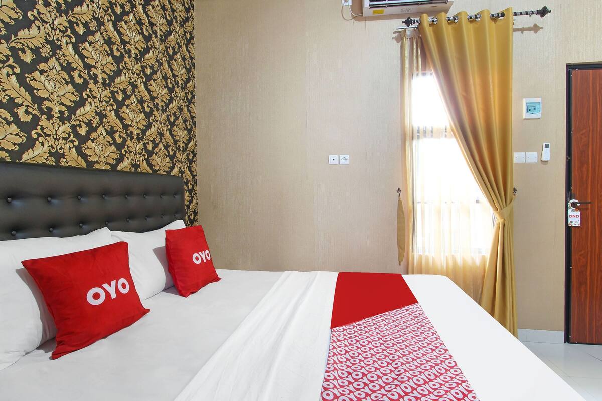 Habitación de hotel con una cama, dos almohadas rojas con la marca "OYO", un plaid estampado, una ventana con cortinas amarillas y una puerta con el letrero "DND".