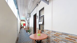 Un étroit couloir extérieur doté d’un sol carrelé, une table ronde sur laquelle se trouve un objet vert et une plante en pot.