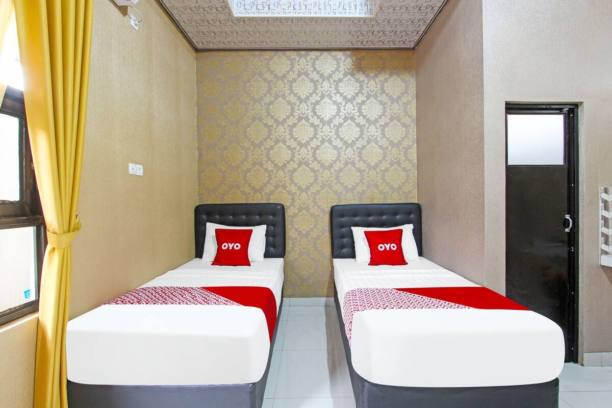 Habitación de hotel con dos camas, cada una con una colcha roja y blanca y un cojín a juego. La habitación tiene papel tapiz estampado y una puerta con un espejo.