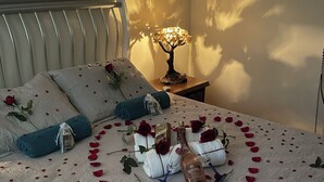 Un lit décoré de pétales de rose disposés en forme de cœur, une bouteille de liqueur, des oreillers et une lampe de chevet.