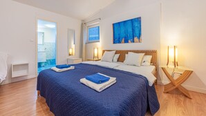 Ein Doppelbett mit blauer Bettdecke, zwei Nachttische mit Lampen, ein Holz-Kopfschild, ein Gemälde und ein durch eine geöffnete Tür einsehbares Badezimmer.