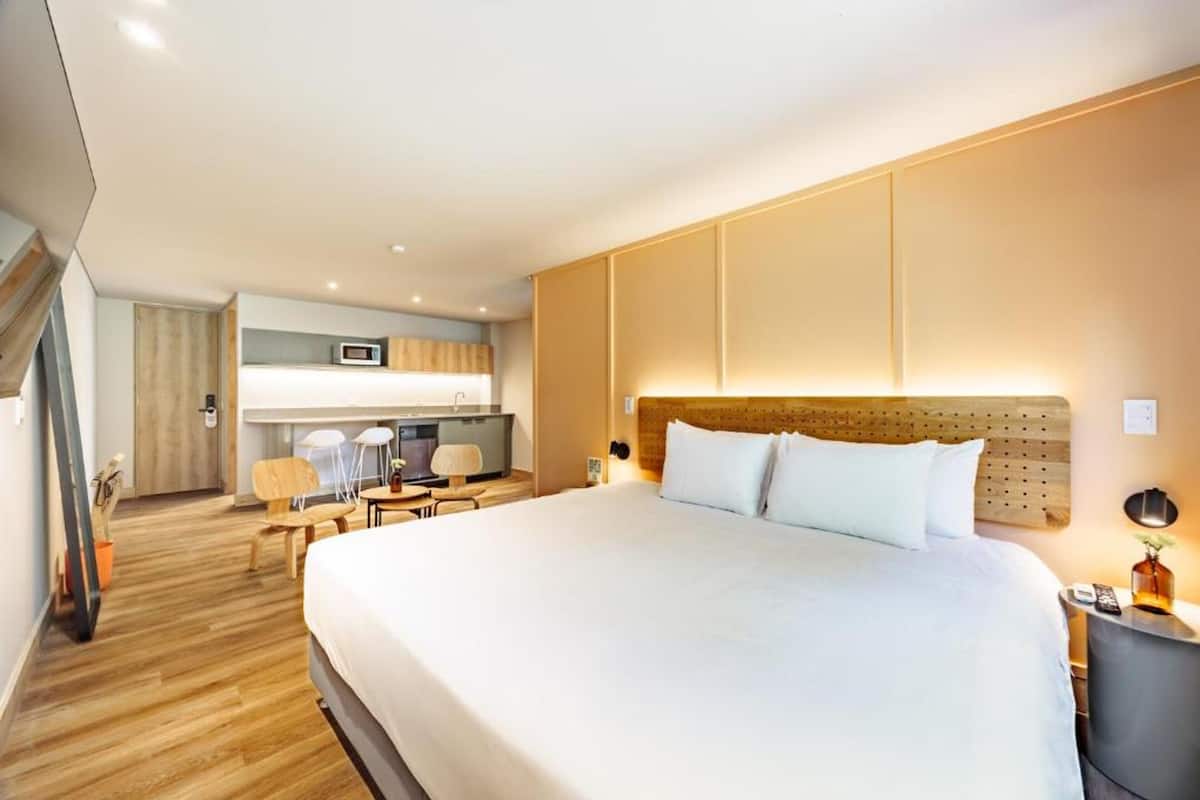 A modern hotel room with a large bed, a desk, a chair, a microwave, and a small kitchenette.