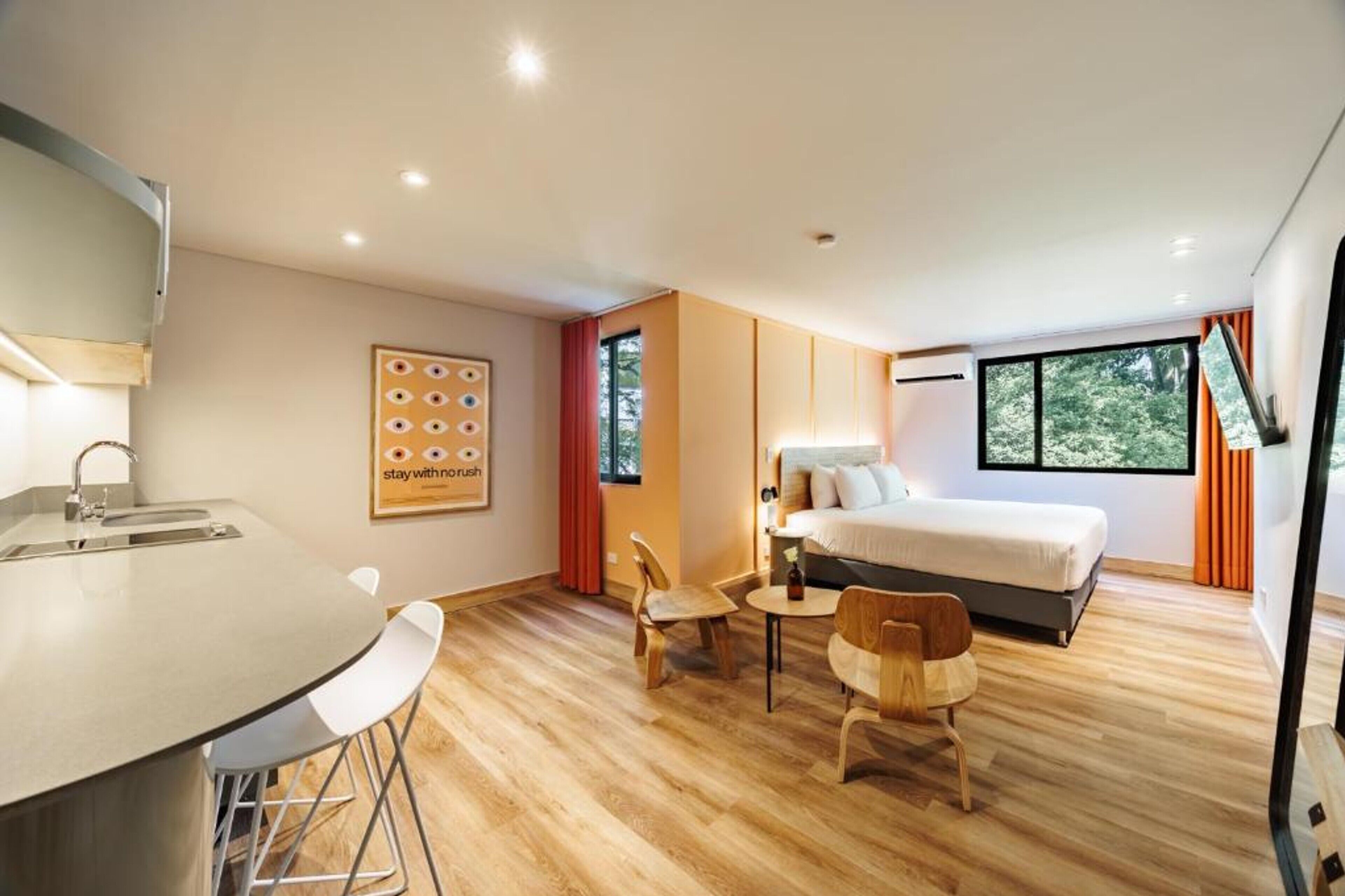 A modern hotel room with a bed, a desk with a chair, a small table with a chair, and a large window with curtains.