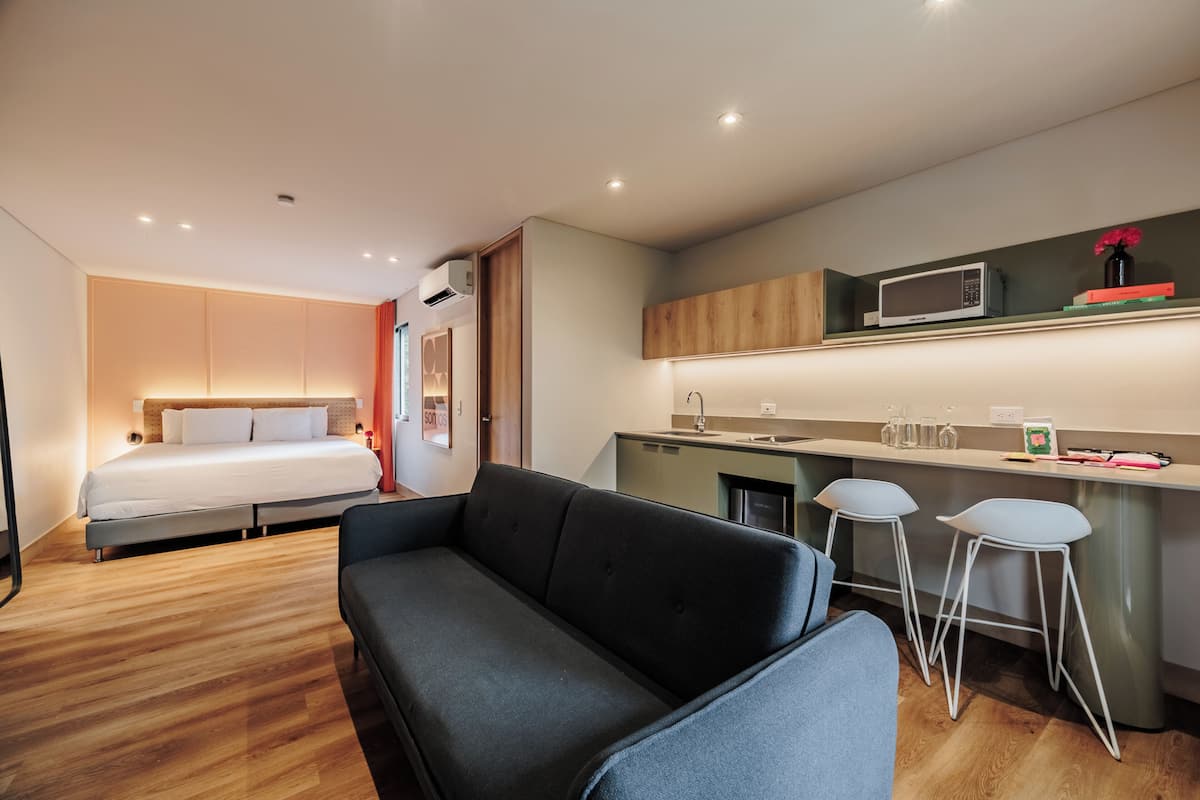 A compact hotel room with a bed, a sofa, a kitchenette, and a small dining area.