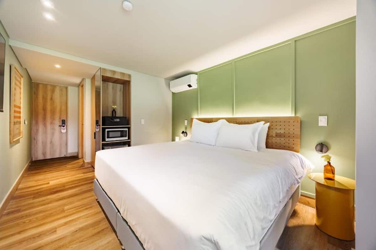 A hotel room with a large bed, a wooden floor, a green accent wall, and a small side table with a lamp.