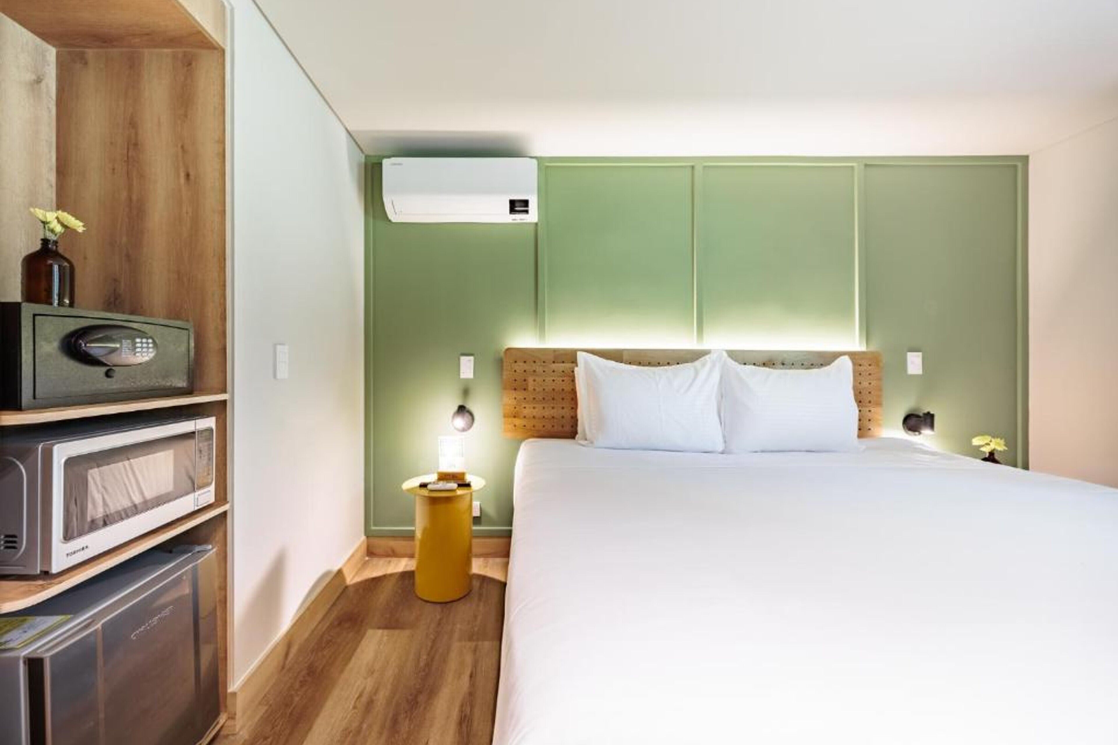 Una habitació d'hotel amb un llit, una taula de nit amb una làmpada, un microones, un frigorífic i un aparell d'aire condicionat.
