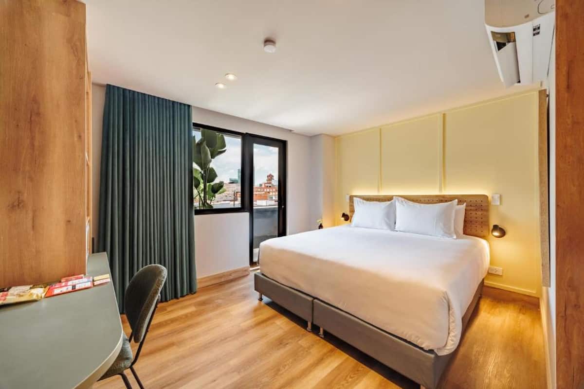 A modern hotel room with a large bed, a desk, and a view of the city through the window.