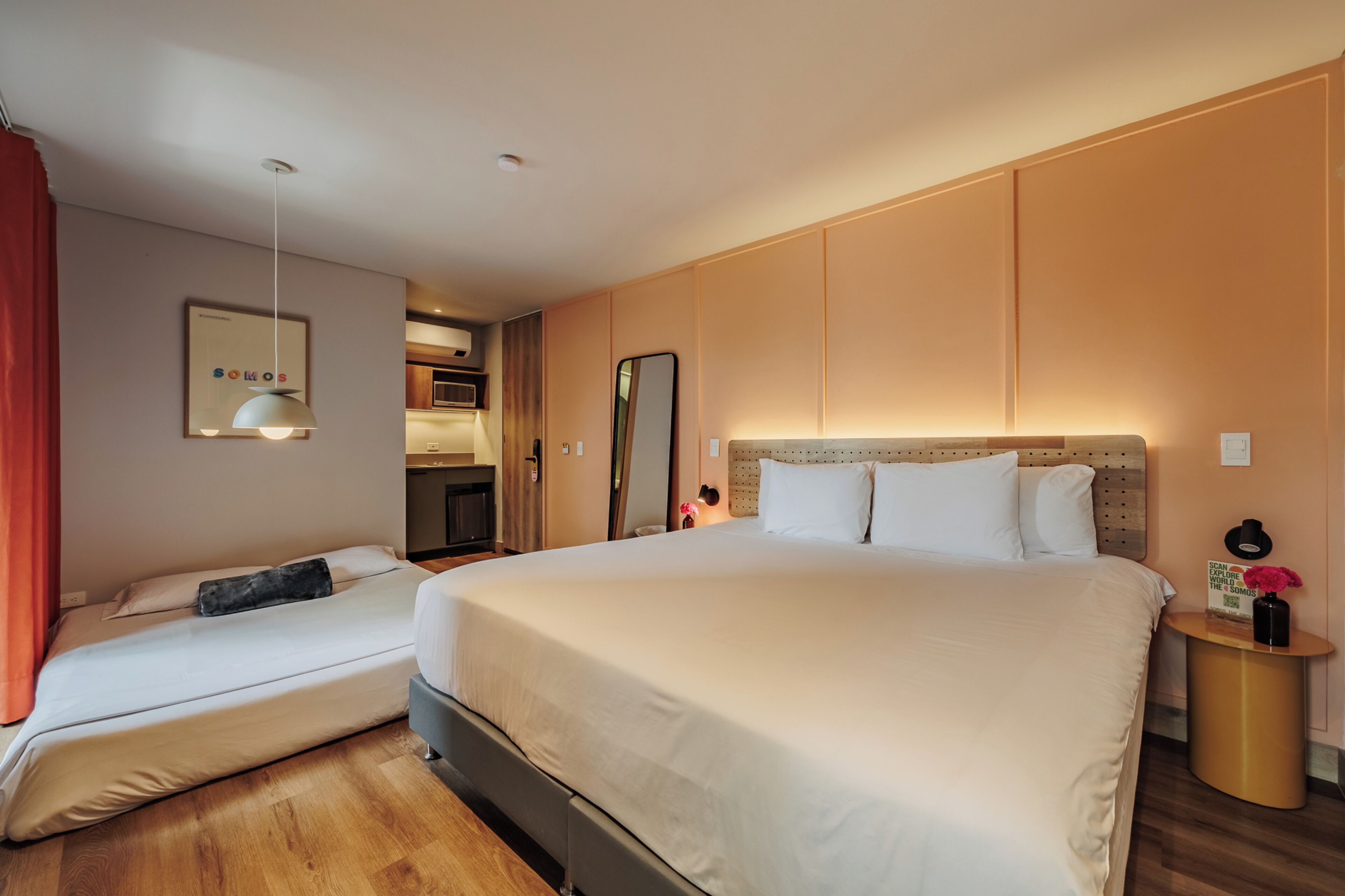 A hotel room with a large bed, a smaller bed, a bedside table with a lamp, a mirror, and a view of the kitchen.