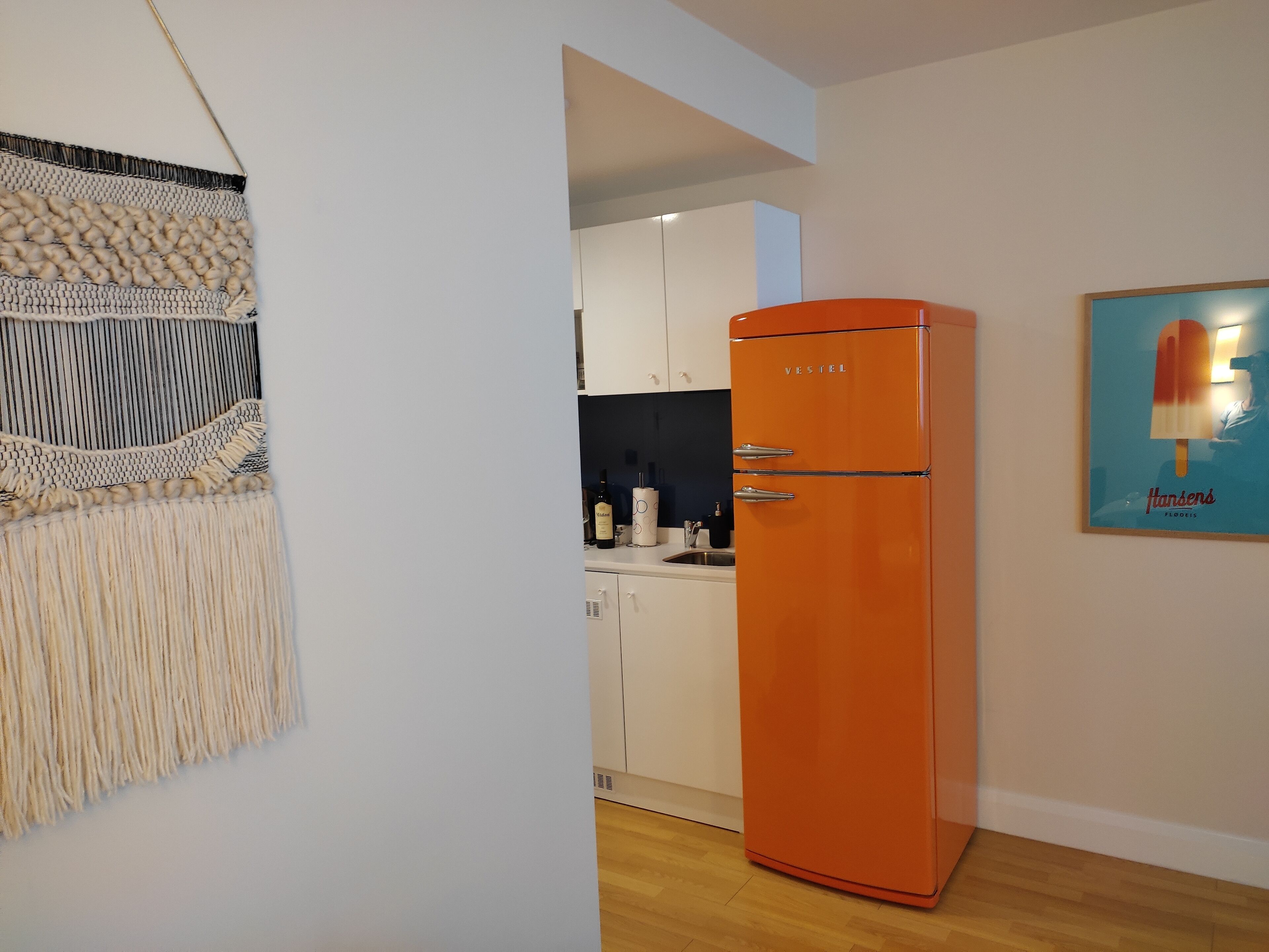 A modern kitchen with a vintage orange refrigerator, white cabinets, and a macrame wall hanging.