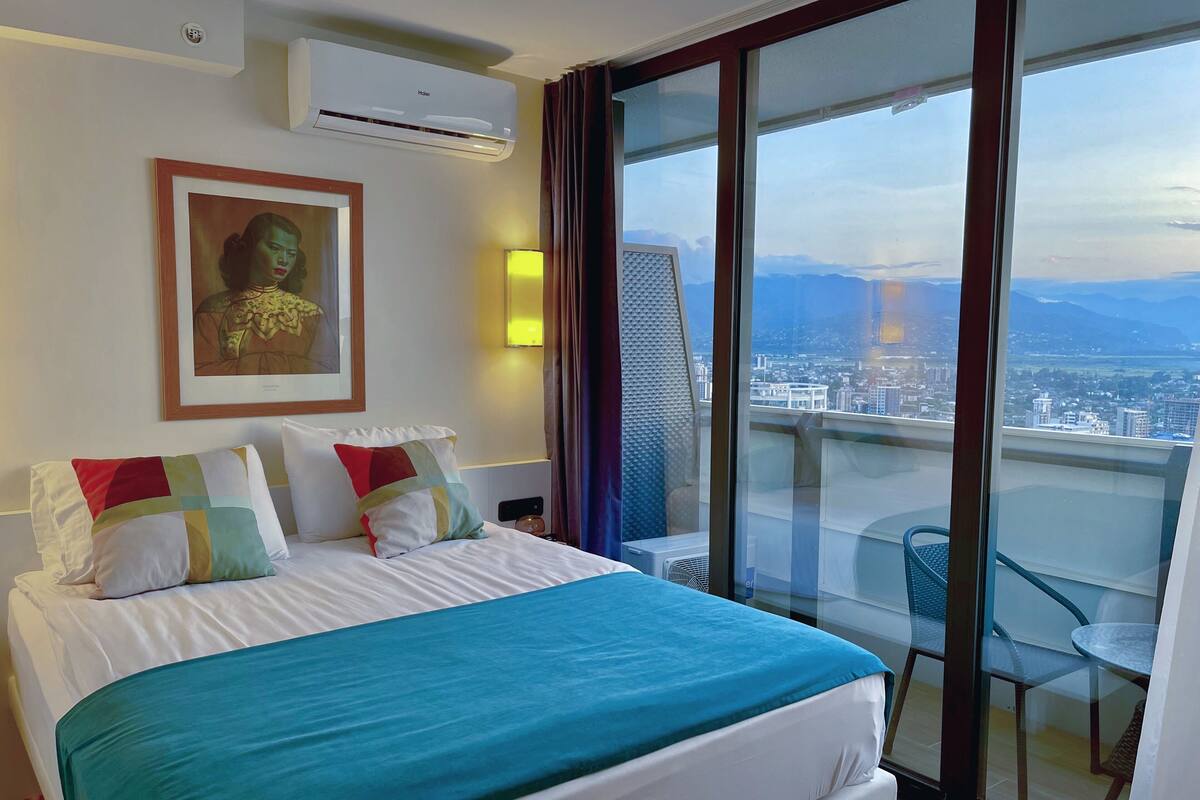 A hotel room with a large bed, a framed artwork on the wall, a city view through the balcony, and an air conditioning unit.