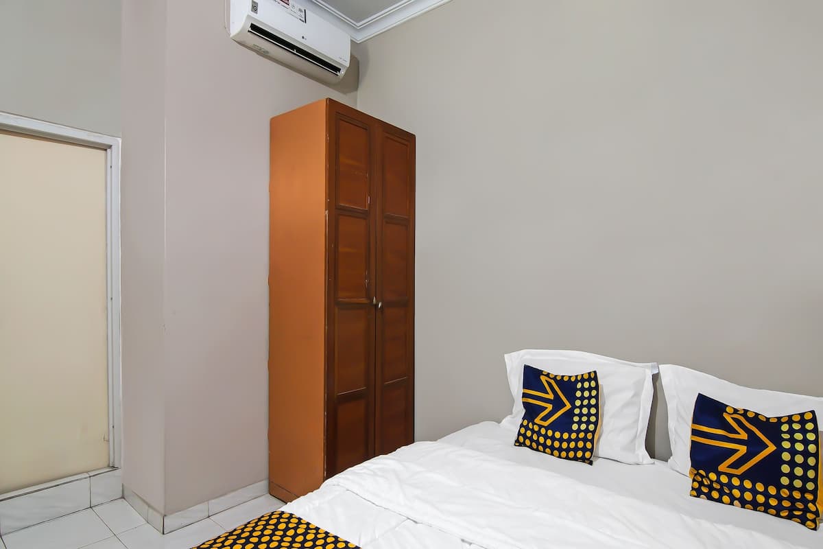 Une chambre avec un lit blanc, deux oreillers à motifs géométriques, une armoire en bois et un climatiseur fixé au mur.