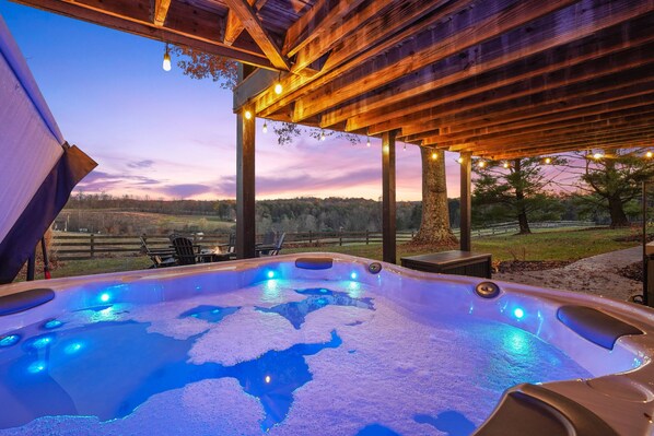 A hot tub under a wooden pergola with string lights, surrounded by a grassy area and trees.