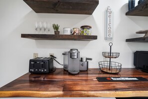 A kitchen counter with a Keurig coffee maker, a toaster, and a wire basket. There are shelves above holding mugs, a plant, and jars. A 'welcome' sign is on the wall.