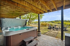 A hot tub under a wooden pergola with a stone patio, outdoor seating, and a view of a grassy landscape. - The Lodge at Shaw Ranch | Hocking Hills, 30 Acres (Rockbridge)