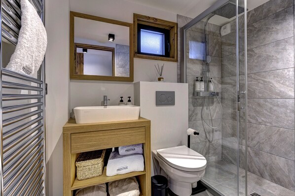 Un baño moderno con lavamanos, tocador y retrete blancos. Hay dos espejos sobre el lavamanos, una ducha con puertas de cristal y un perchero con toallas.