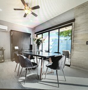 Un espace de salle à manger moderne, doté d’une grande table, de chaises noires, d’un ventilateur de plafond et offrant une vue sur la verdure grâce à de grandes fenêtres.