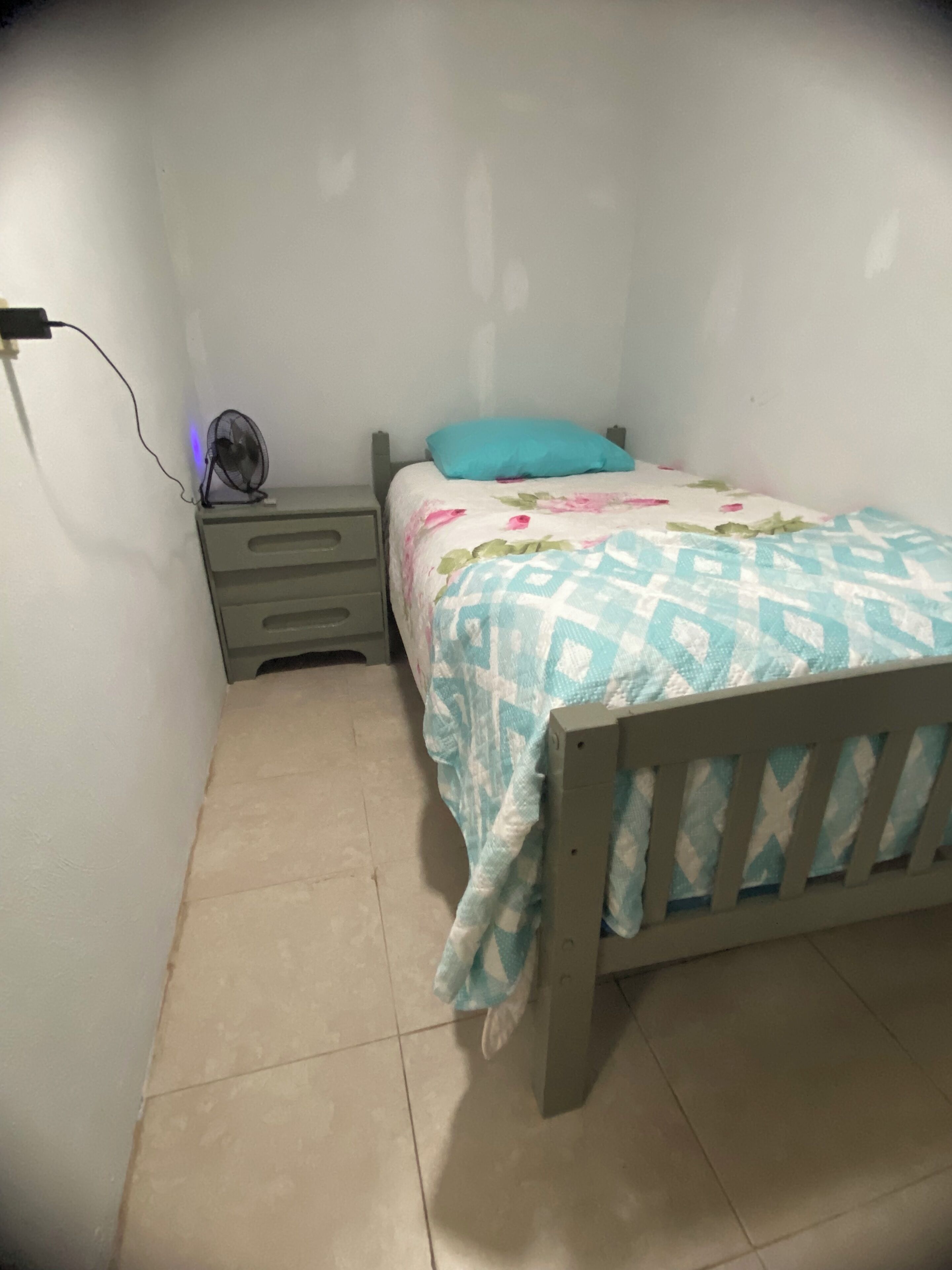 A small, single-bed room with a wooden headboard, a bedside table with drawers, a wall-mounted fan, and a bed with a floral-patterned bedspread.