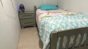 A small, single-bed room with a wooden headboard, a bedside table with drawers, a wall-mounted fan, and a bed with a floral-patterned bedspread.