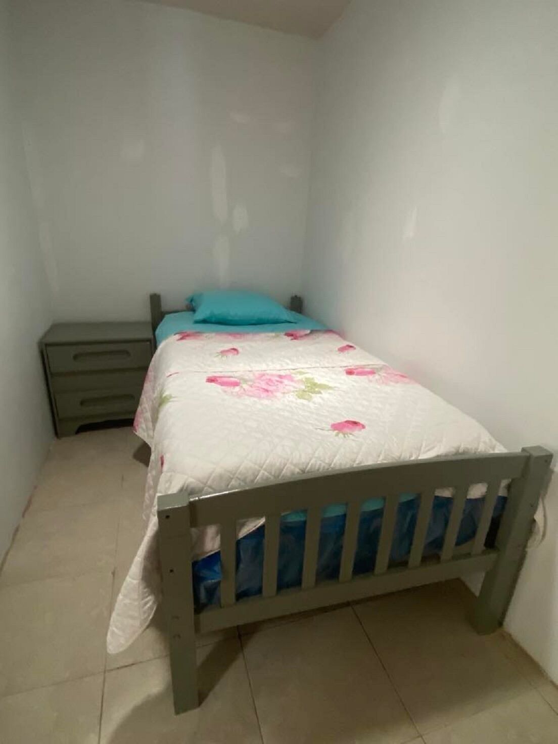 Una habitación pequeña con cama individual, cabecera de madera, mesita de noche y colcha estampada con motivos florales.