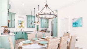 A modern kitchen with teal cabinets, a central island, and a dining area with wicker chairs and a round table. - Pura Vida Villa: Luxury Coastal Retreat With Private Heated Pool (Murrells Inlet)