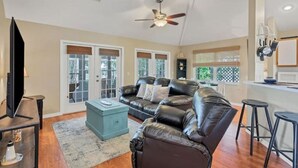 A living room with a black leather sofa, a coffee table, and a TV. There is a ceiling fan, a window with blinds, and a kitchen area visible through the window. - Long-Term Discount!Unique 3BR canal home, Private Dock, Boat Lift, Deck! (Punta Gorda)