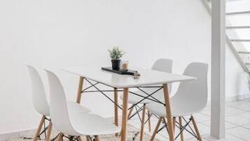 Un espace repas moderne avec une table et des chaises blanches, une horloge au mur et un escalier en arrière-plan.
