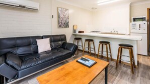 Una sala moderna con un sofá de cuero negro, una mesa de centro de madera y una cocina con encimera blanca y taburetes.