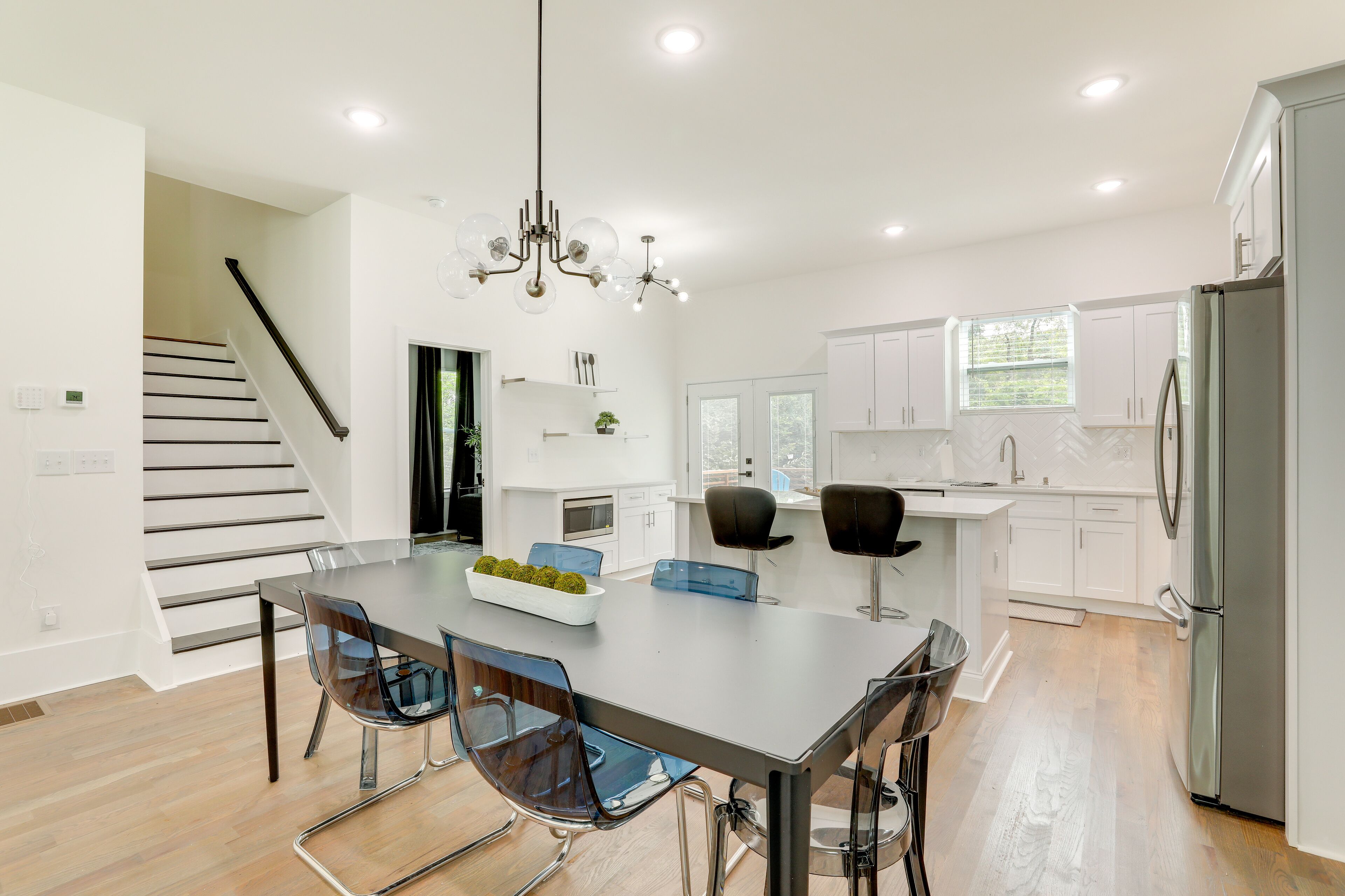 Una cocina moderna con armarios blancos, un refrigerador de acero inoxidable y un comedor con mesa y sillas de vidrio.