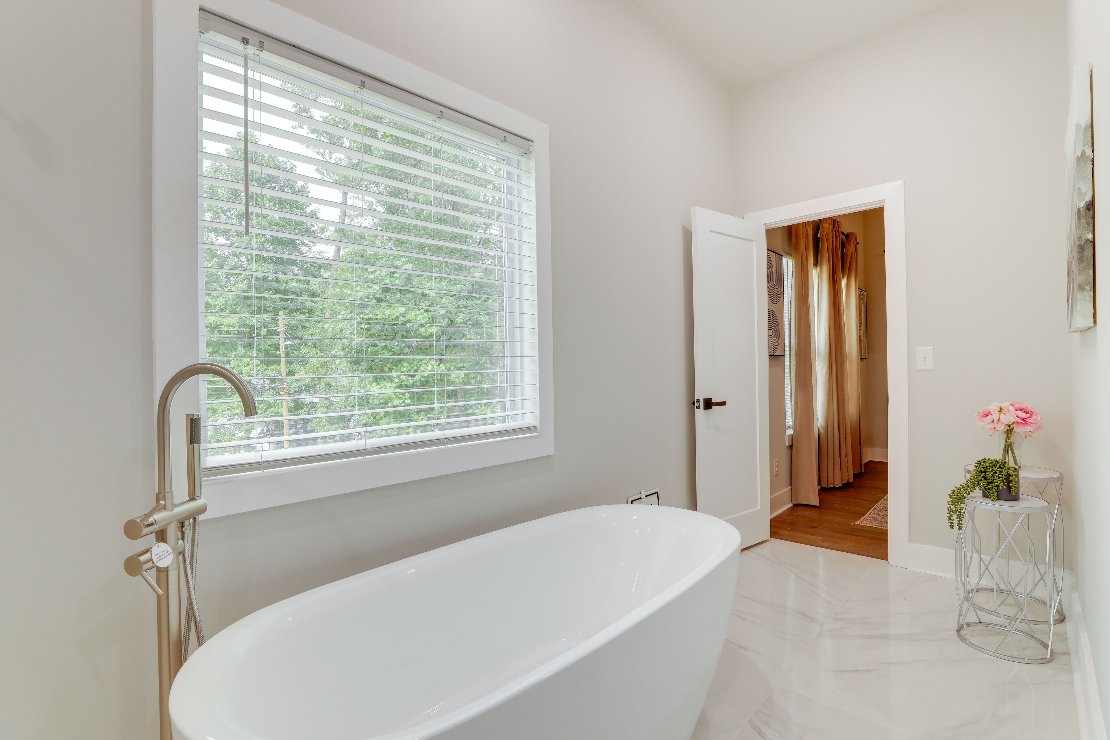 Une salle de bain moderne avec une baignoire sur pieds, une fenêtre avec des stores et une porte donnant sur une autre pièce.