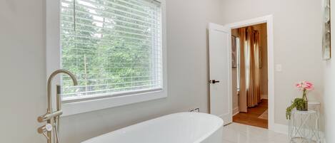 Une salle de bain moderne avec une baignoire sur pieds, une fenêtre avec des stores et une porte donnant sur une autre pièce.