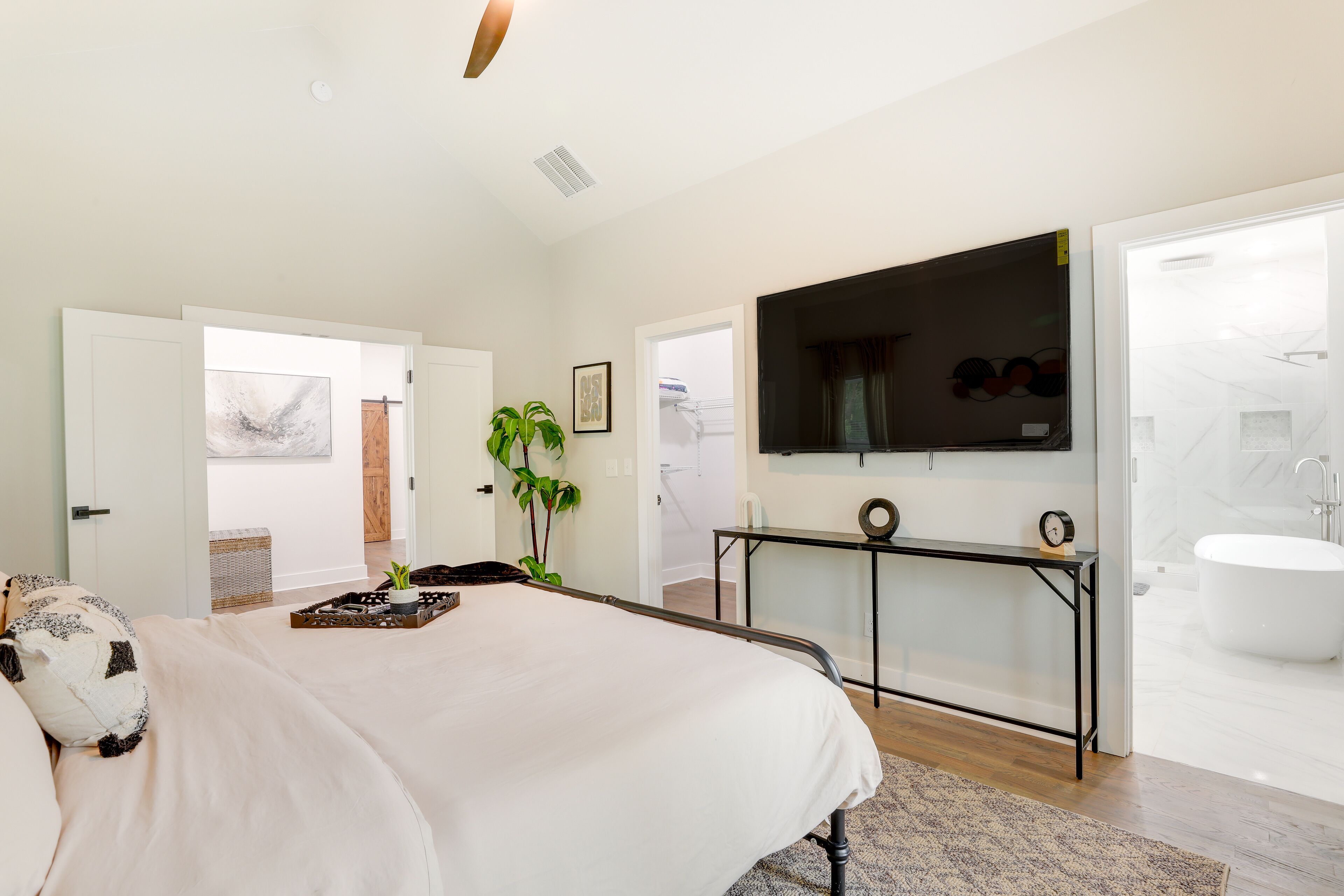 A bedroom with a large bed, a TV mounted on the wall, a bedside table, and a bathroom with a bathtub and toilet visible through an open door.