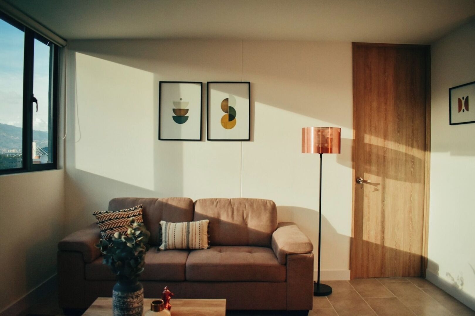 Un salon moderne avec un canapé, une table basse et un lampadaire. Deux œuvres d’art encadrées ornent le mur, et on peut voir un pont à travers la fenêtre.