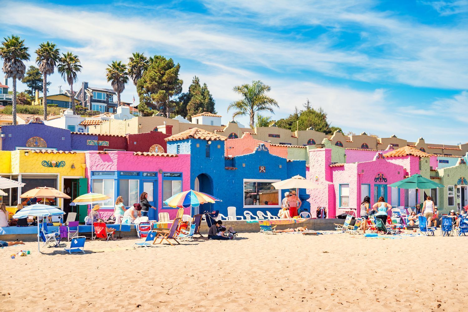 Une plage avec des bâtiments aux couleurs vives, des chaises de plage, des parasols et des gens.
