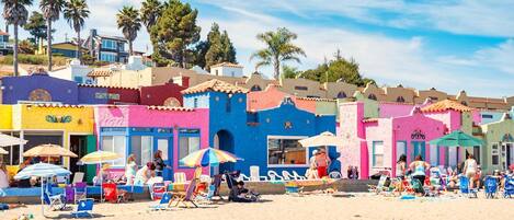 Une plage avec des bâtiments aux couleurs vives, des chaises de plage, des parasols et des gens.