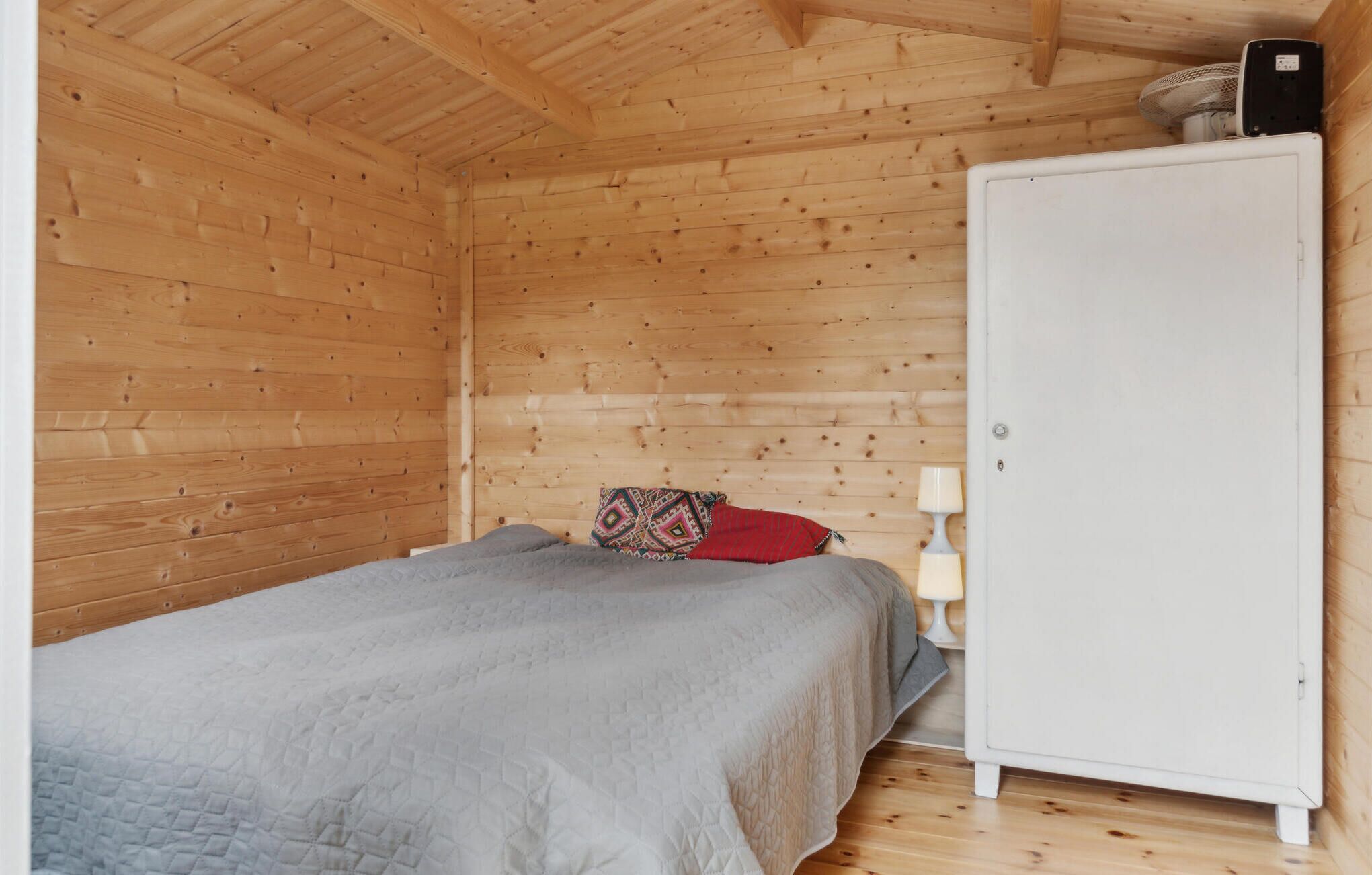 A small, cozy room with wooden walls and a single bed. There is a white wardrobe and a small table with a lamp.