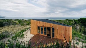 Een modern houten gebouw met grote glazen deuren, gelegen op een zandig terrein met uitzicht op zee en verre bergen.