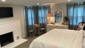 Une chambre avec une cheminée en briques blanches, un grand lit, une table de chevet avec une lampe, un miroir et une chaise.